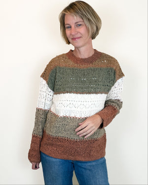 A woman wearing a chunky knit crewneck sweater with horizontal stripes in rust brown, olive green, and white, paired with blue jeans, standing against a plain off-white wall.