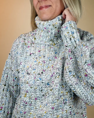 A close-up of a woman wearing a chunky knit turtleneck sweater with a heathered light gray base and multicolored yarn flecks, set against a solid beige background.