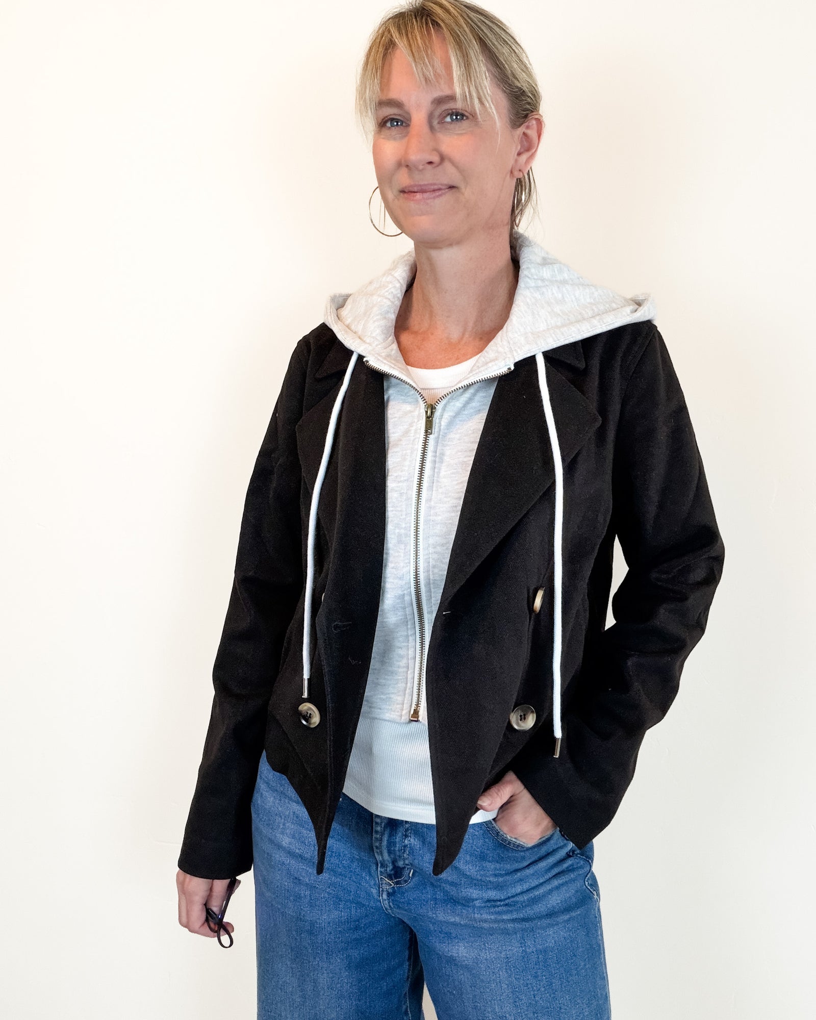 A woman wearing a black suede-look hooded jacket over a light gray zip-up hoodie and blue jeans, standing against a plain off-white background.