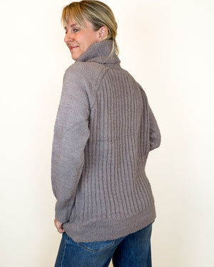 A woman wearing a gray ribbed turtleneck sweater, shown from a three-quarter back view against a plain off-white background.