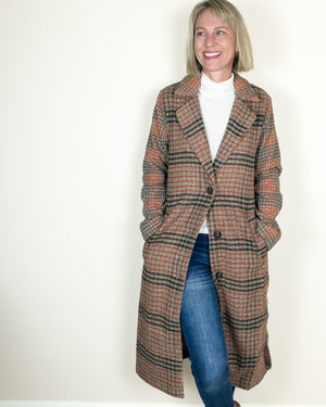 A woman modeling a long, double-breasted plaid coat in brown, beige, and black, worn over a white turtleneck and blue jeans against a plain white wall.