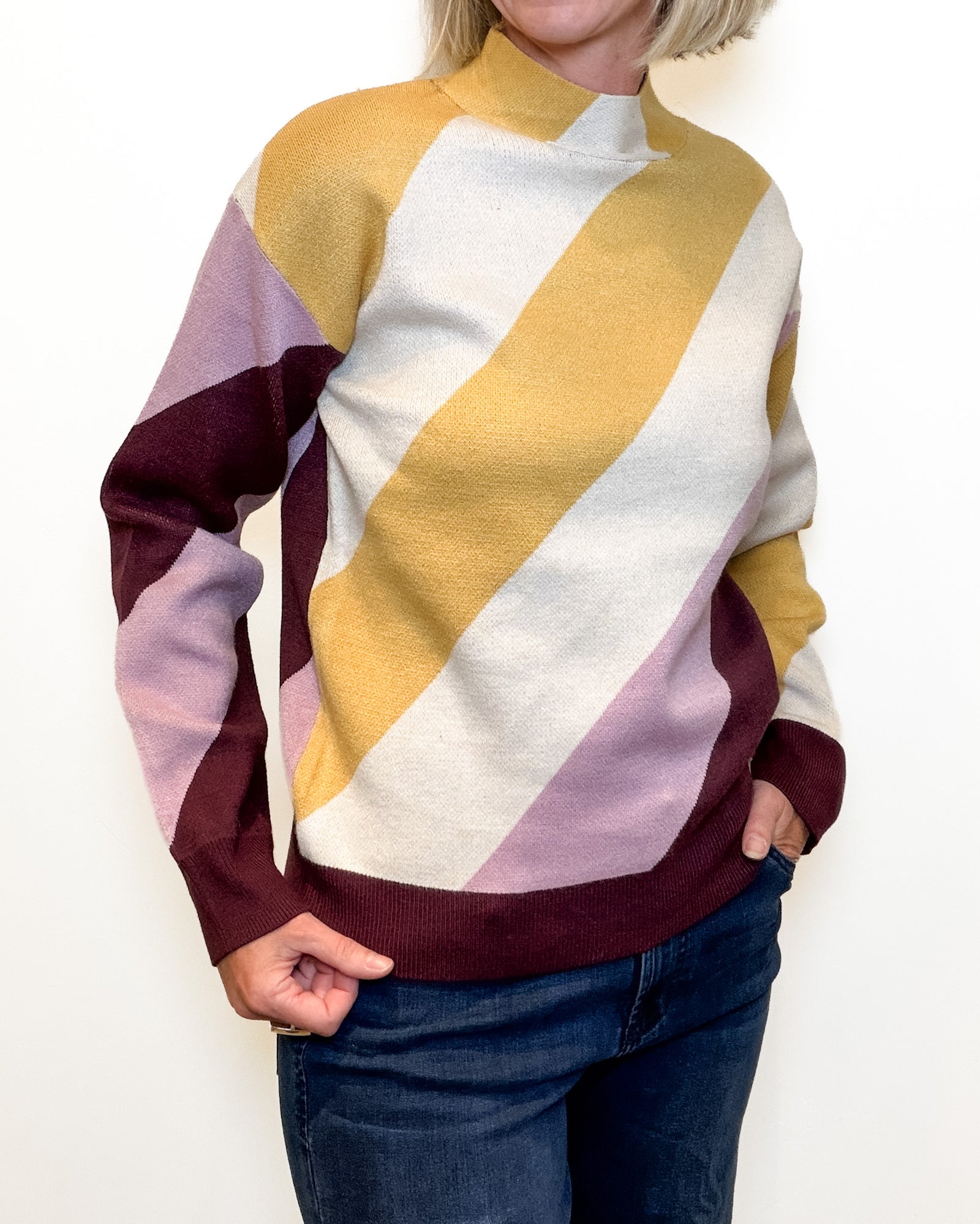 A person wearing a color-block knit sweater with diagonal stripes in mustard yellow, cream, dusty lavender, and burgundy, paired with dark blue jeans against a plain white background.