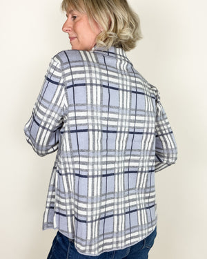 A woman shown from the back wearing a light blue, white, and navy plaid flannel shirt and blue jeans, standing against a plain off-white background.