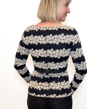 A woman shown from the back wearing a long-sleeve knit sweater with horizontal black and cream lace-like stripes, paired with black pants, against a plain white background.