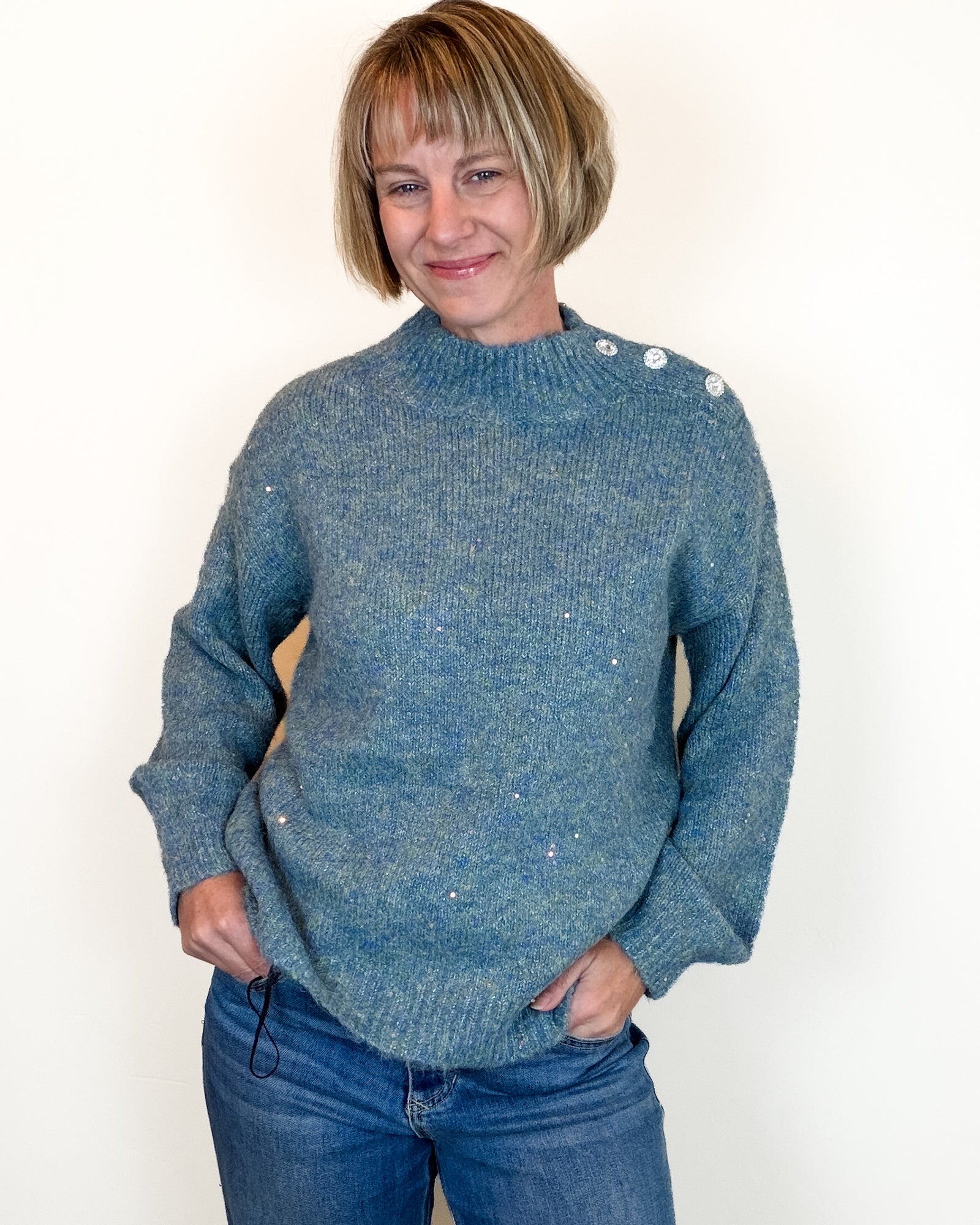 A close-up of a person wearing a textured blue knit sweater with three decorative silver buttons at the collar and small scattered sequins.