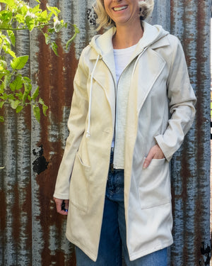 A woman wearing a cream-colored hooded jacket with a front zipper and drawstrings, standing in front of a corrugated metal fence.