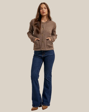 A woman modeling a cropped taupe-brown knit cardigan with front buttons and patch pockets, paired with dark blue flared jeans and tan pointed-toe heels, against a plain light beige studio background.