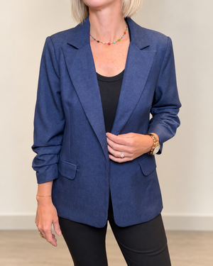 A woman wearing a blue textured blazer over a black top and black pants, standing against a neutral beige wall.