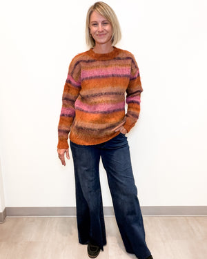 A woman modeling a fuzzy, long-sleeve crewneck sweater with horizontal stripes in orange, brown, pink, and purple, paired with dark blue wide-leg jeans.