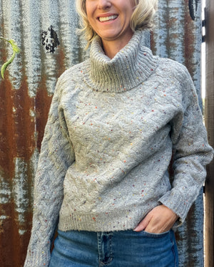A woman wearing a light gray cable-knit turtleneck sweater with subtle multicolor flecks, paired with blue jeans, standing in front of a rustic corrugated metal wall.