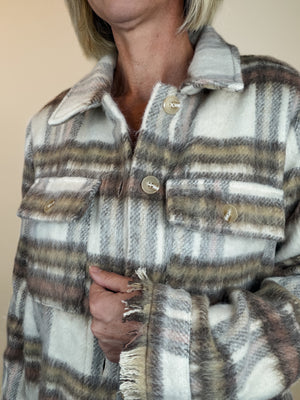 A close-up of a person wearing a soft plaid button-front jacket or overshirt in off-white, beige, brown, and muted gray tones. The fabric has a fuzzy texture, frayed edges, and large buttons.