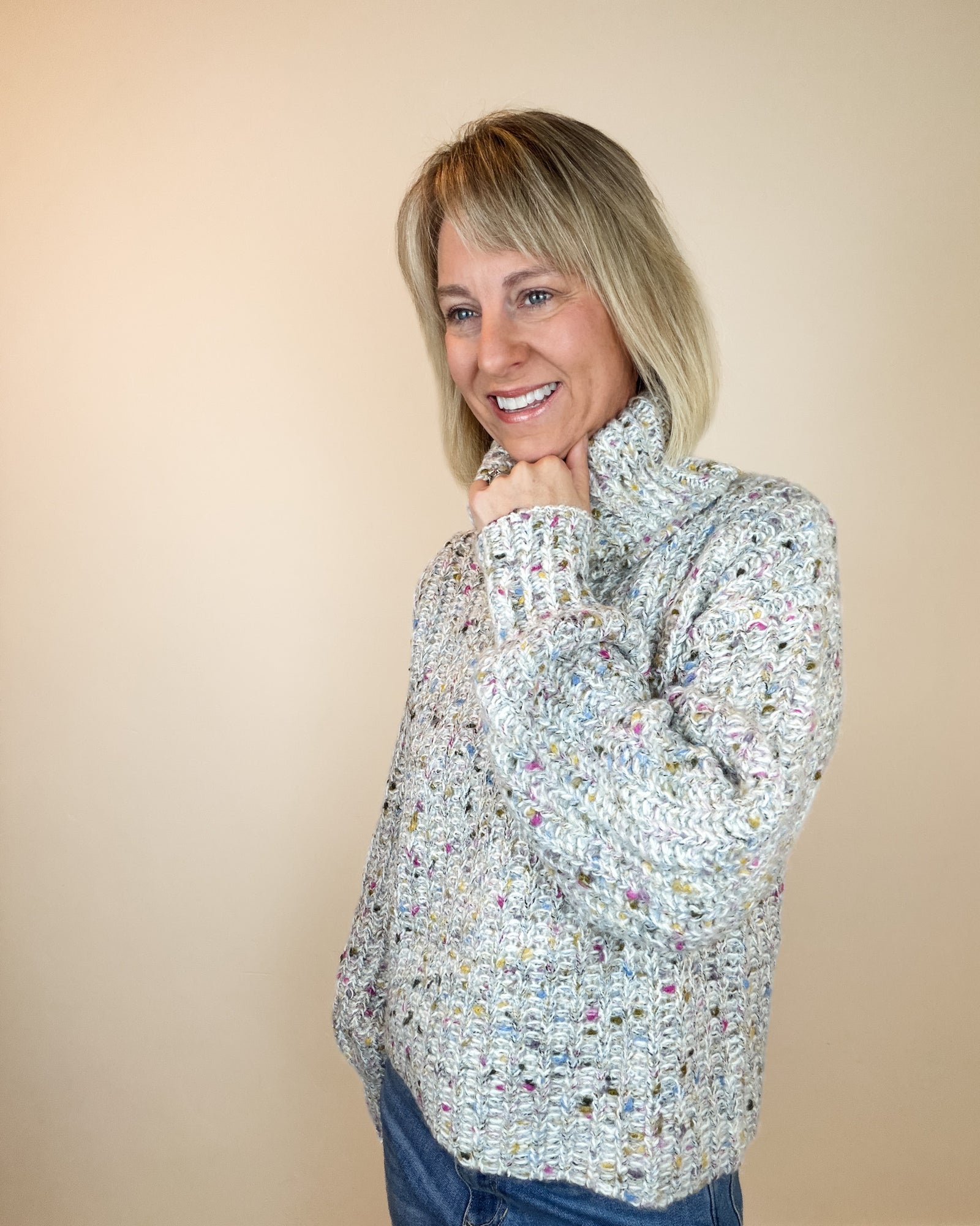 A smiling blonde woman wearing a chunky knit turtleneck sweater with a multicolor variegated pattern, paired with blue jeans, standing against a plain off-white wall.