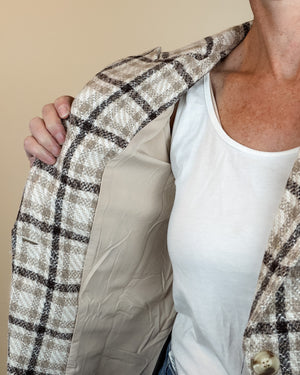 A close-up of a person wearing an open plaid blazer over a white tank top, with the blazer's beige and brown patterned fabric visible against a plain beige background.