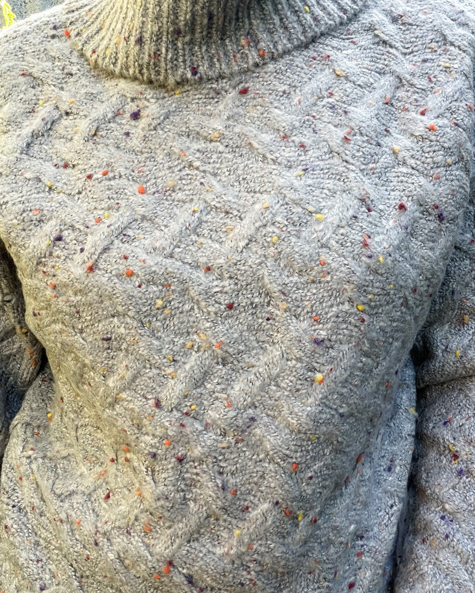 A woman wearing a chunky cable-knit turtleneck sweater in a heathered oatmeal gray with tiny multicolor flecks, paired with blue jeans, standing against a rustic corrugated metal wall.