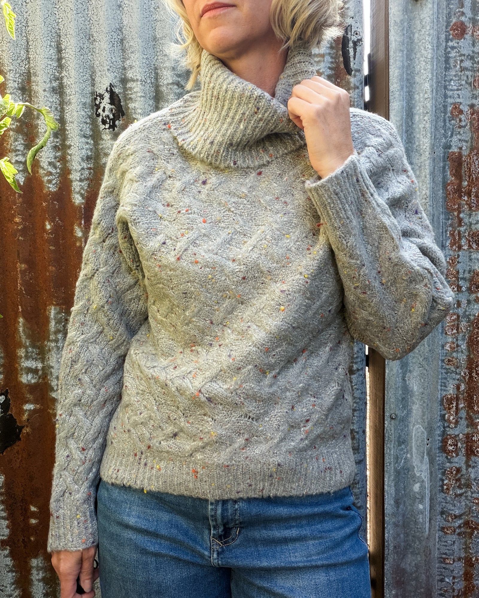 A woman wearing a chunky cable-knit turtleneck sweater in a heathered oatmeal gray with tiny multicolor flecks, paired with blue jeans, standing against a rustic corrugated metal wall.