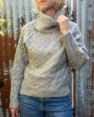 A woman wearing a chunky cable-knit turtleneck sweater in a heathered oatmeal gray with tiny multicolor flecks, paired with blue jeans, standing against a rustic corrugated metal wall.