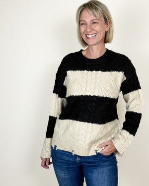 A smiling woman with short blonde hair wearing a black and cream cable-knit sweater and blue jeans, standing against a plain off-white background.