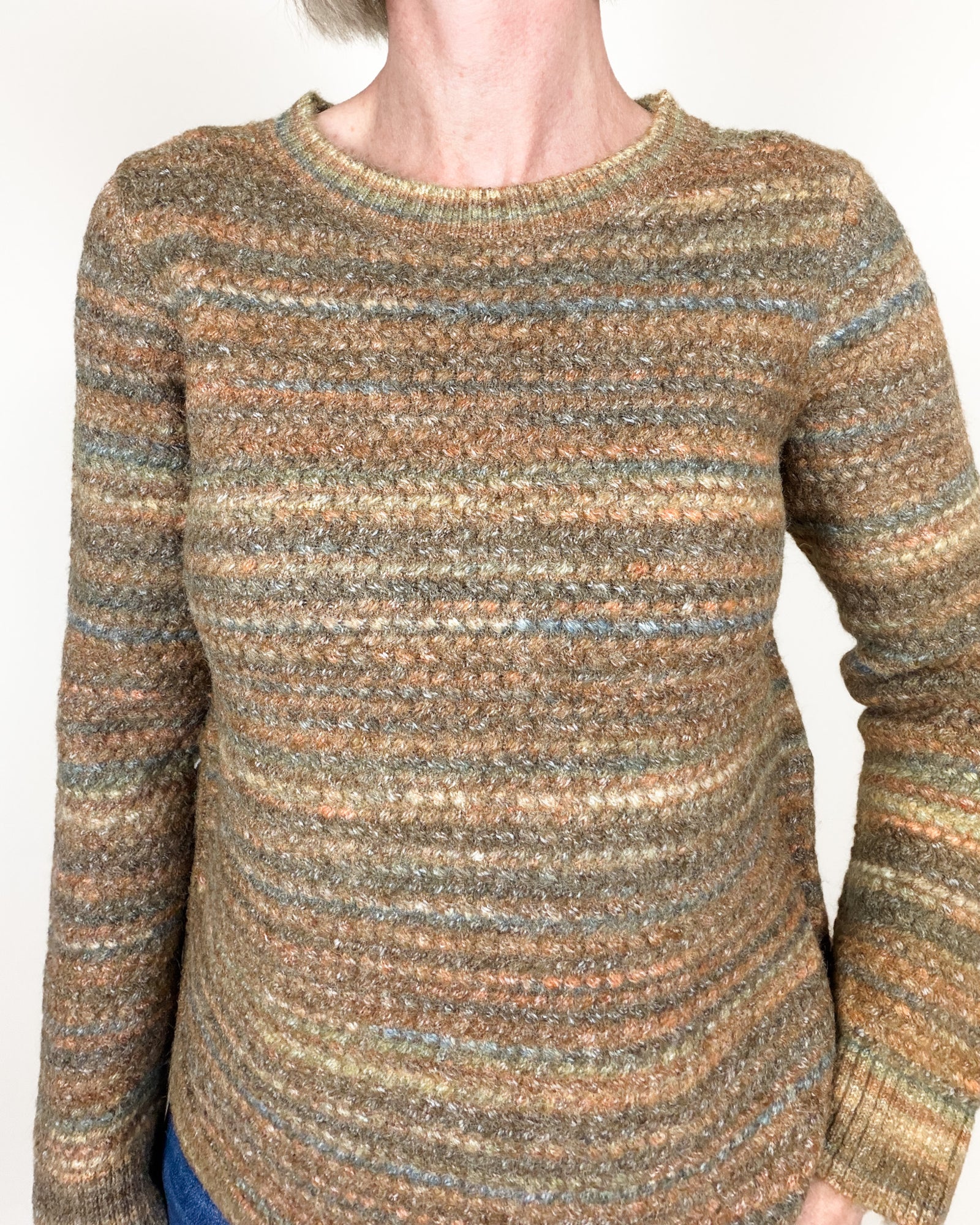 A woman with short blonde hair wearing a textured, striped knit sweater in earthy brown, beige, and muted blue tones, paired with blue jeans, standing against a plain white wall.