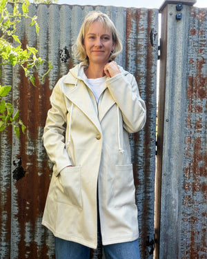 A woman wearing a cream-colored hooded jacket with a drawstring and front pockets, standing in front of a weathered corrugated metal fence.