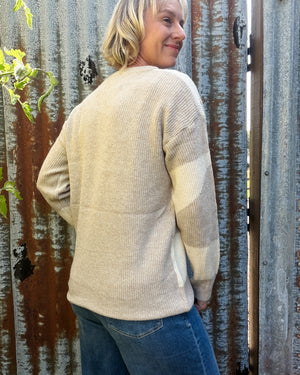 A woman wearing a cream ribbed knit sweater with beige color-blocked panels on the sleeves, standing in front of a weathered corrugated metal fence.