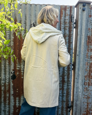 A person viewed from behind wearing a light cream hooded jacket, standing in front of a weathered corrugated metal fence.