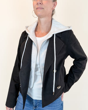 A woman wearing a black zip-front jacket layered over a light gray hooded sweatshirt and white T-shirt, paired with blue jeans, against a plain off-white background.