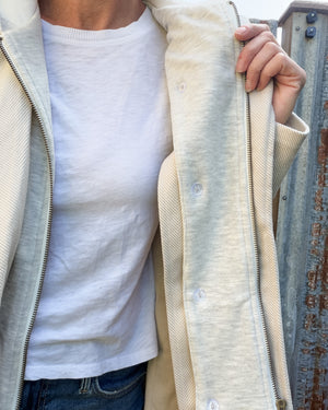 A close-up of a person wearing a cream-colored zip-up hoodie over a white t-shirt and blue jeans, standing next to a weathered metal structure.