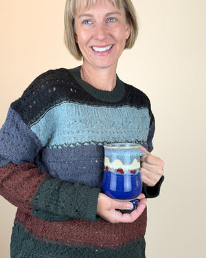 A smiling woman with short blonde hair wearing a chunky, multi-colored knit sweater holds a blue ceramic mug with a striped pattern. The sweater features horizontal bands of black, light blue, slate blue, dark green, and rust brown.