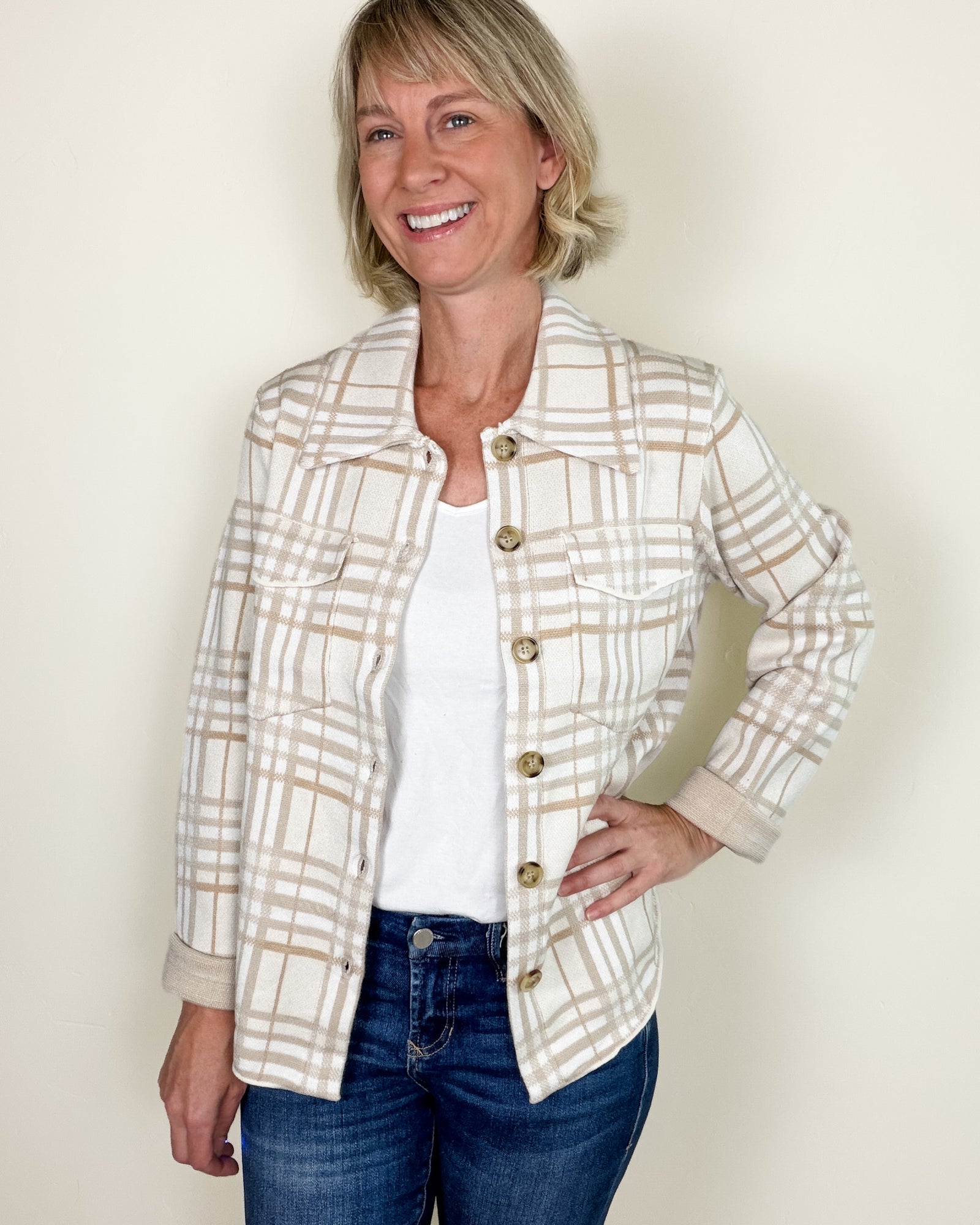 A smiling woman wearing a cream and tan plaid button-front jacket over a white top and blue jeans, standing against a plain light beige wall.