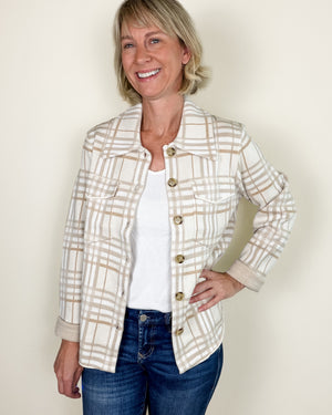 A smiling woman wearing a cream and tan plaid button-front jacket over a white top and blue jeans, standing against a plain light beige wall.