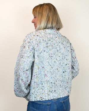 A woman with blonde hair is shown from the back wearing a chunky knit cardigan with a multicolor variegated pattern on a light blue-gray base, paired with blue jeans, against a plain off-white background.