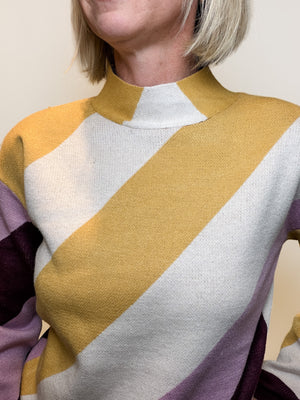 A close-up of a person wearing a knit mock-neck sweater with bold diagonal color-block stripes in mustard yellow, off-white, and lavender purple.