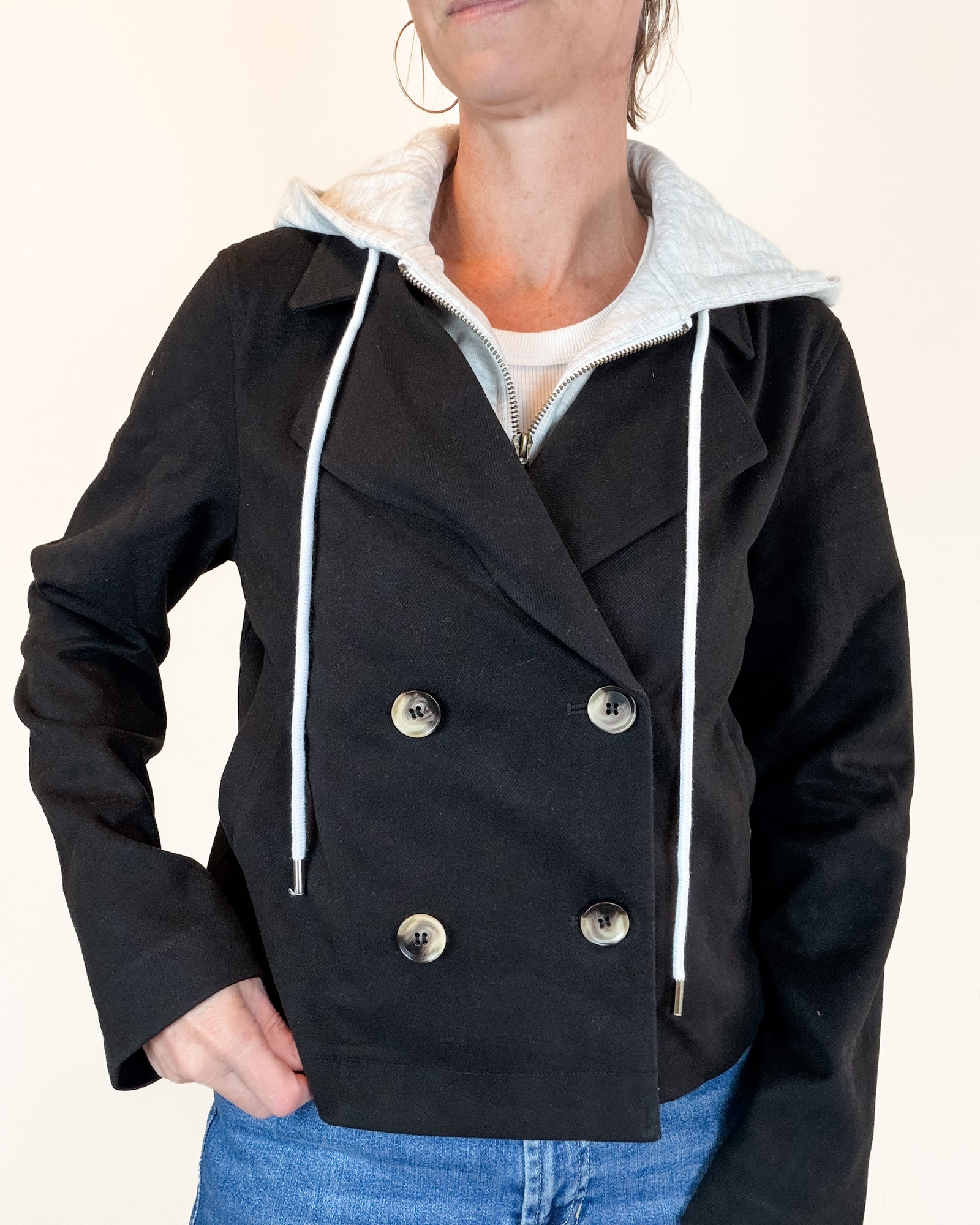 A woman wearing a black suede-look hooded jacket over a light gray zip-up hoodie and blue jeans, standing against a plain off-white background.