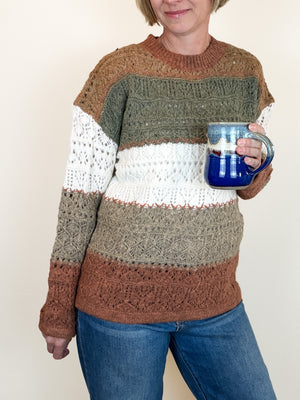 A person wearing a chunky knit striped sweater in brown, olive green, white, and taupe, holding a blue ceramic mug against a plain beige background.