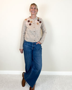 A woman stands indoors wearing a light beige button-front cardigan with colorful floral embroidery across the chest, blue wide-leg jeans, and brown boots against a plain off-white wall.