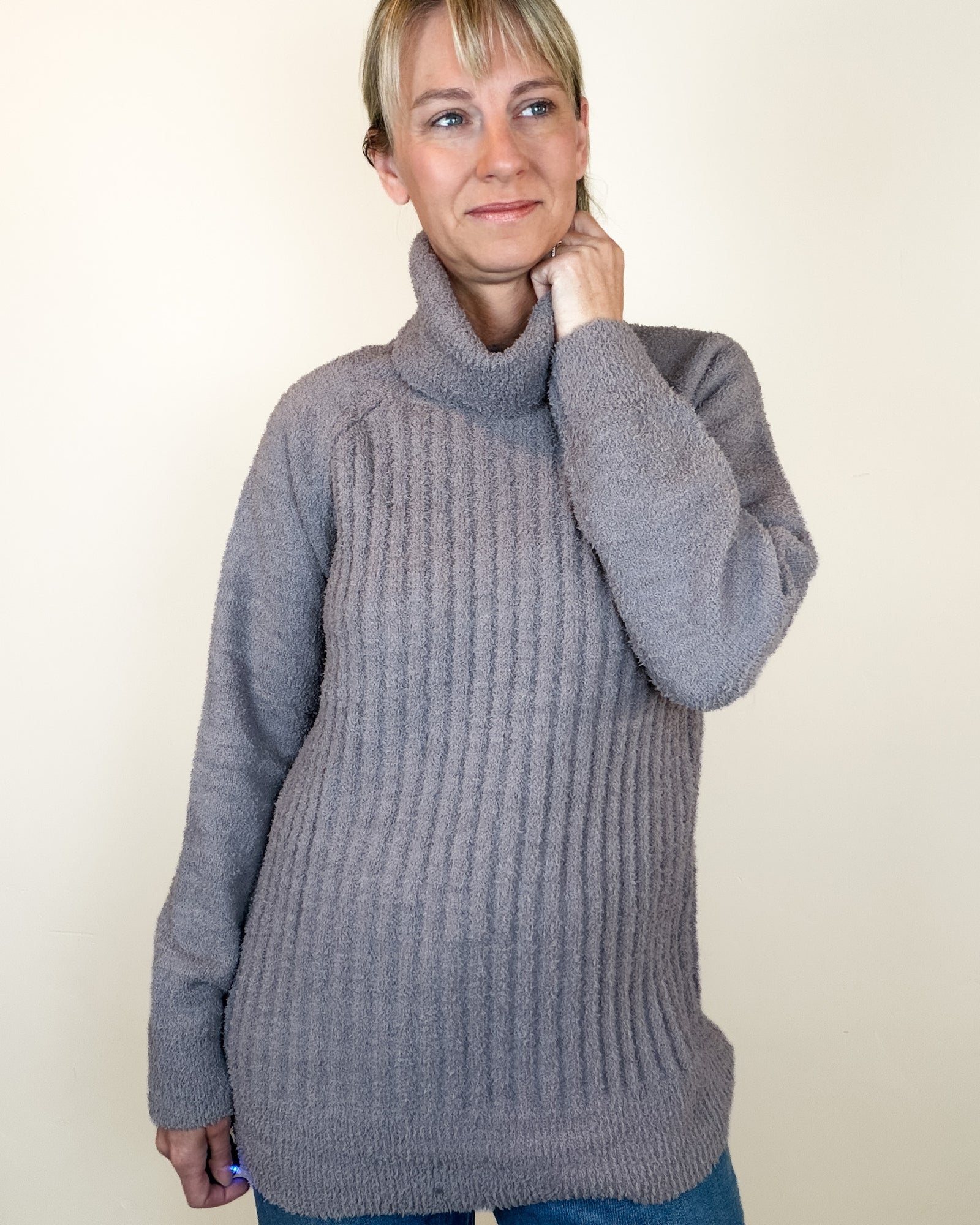 A woman wearing a chunky ribbed turtleneck sweater in a dusty mauve-gray color, paired with blue jeans, standing against a plain off-white background.