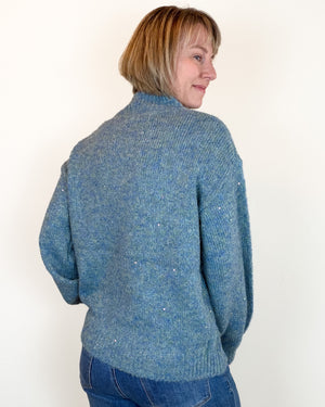 A woman with short blonde hair is shown from the back wearing a textured blue knit sweater and blue jeans against a plain white background.