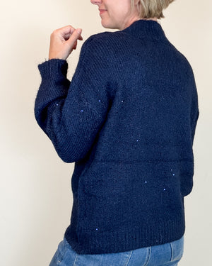 A person shown from the back wearing a dark navy blue ribbed knit sweater with subtle silver speckles, paired with blue jeans, against a plain light beige background.
