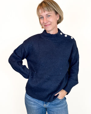 A woman wearing a dark navy blue ribbed knit sweater with silver buttons on the shoulder, paired with blue jeans, against a plain white background.
