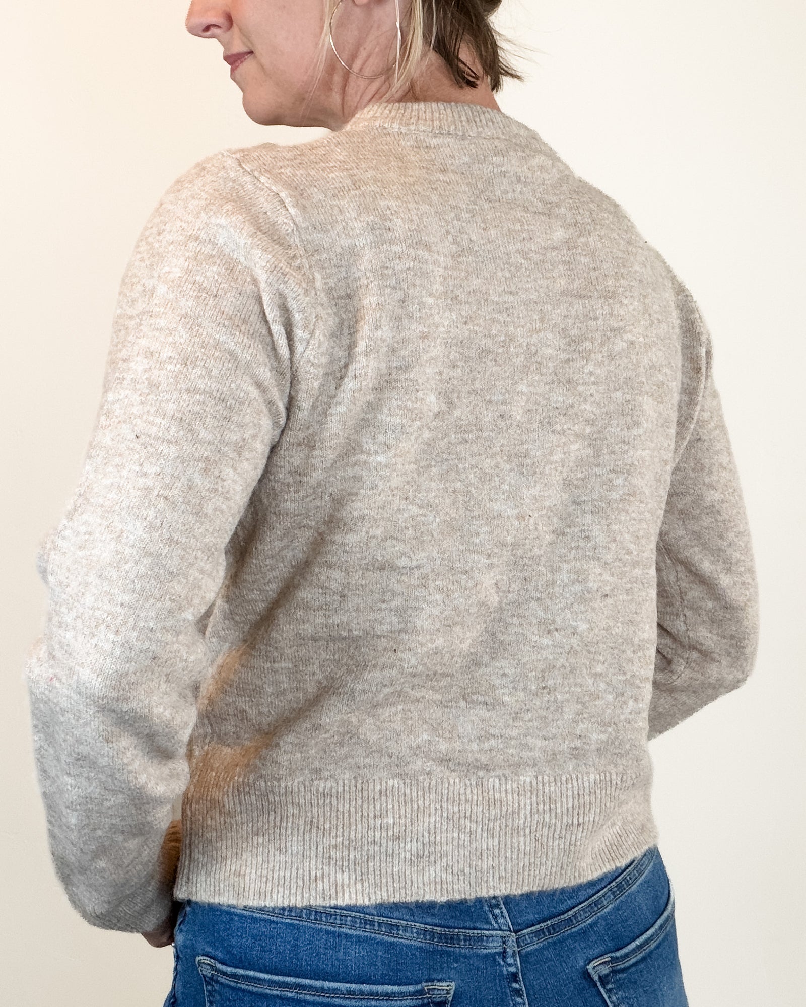 A woman wearing a light beige knit cardigan with brown and pink crocheted flower details on the shoulders, paired with blue jeans, against a plain beige background.