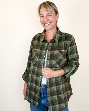 A smiling woman wearing a green and brown plaid button-up shirt over a white undershirt and blue jeans, standing against a plain off-white background.