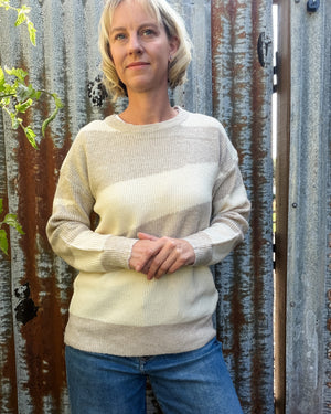 A woman with short blonde hair wearing a cream and beige striped knit sweater and blue jeans, standing in front of a rustic corrugated metal gate.
