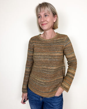 A woman with short blonde hair wearing a textured, striped knit sweater in earthy brown, beige, and muted blue tones, paired with blue jeans, standing against a plain white wall.