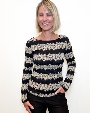 A woman with blonde hair wearing a black and cream patterned knit sweater and black pants, standing against a plain white wall.