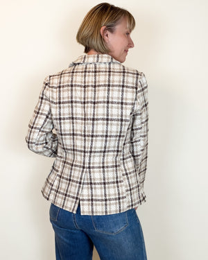 A woman shown from the back wearing a beige and brown plaid blazer with blue jeans, standing against a plain off-white wall.