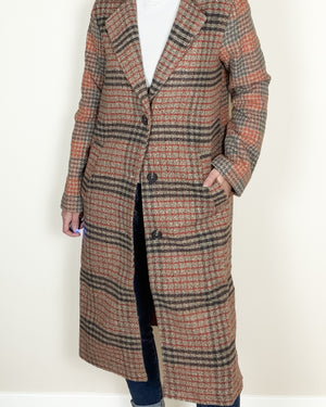 A person wearing a mid-length plaid coat over a white top and dark jeans, standing against a plain off-white wall. The coat has a warm rust-orange, brown, and dark charcoal plaid pattern.