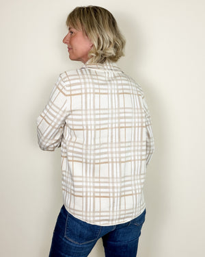 A woman with blonde hair is shown from the back wearing a cream-colored plaid button-down shirt with thin tan stripes and blue jeans, standing against a plain off-white wall.