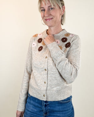 A woman wearing a light beige knit cardigan with brown and pink crocheted flower details on the shoulders, paired with blue jeans, against a plain beige background.