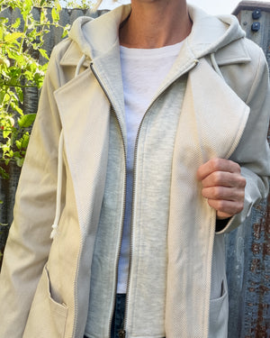 A person wearing a light beige hooded zip-front jacket over a white t-shirt, photographed outdoors against a rustic wooden fence and greenery.