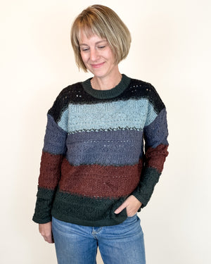 A woman wearing a multi-colored knit sweater with horizontal stripes in black, light blue, slate blue, burgundy, and dark green, paired with blue jeans, against a plain off-white background.